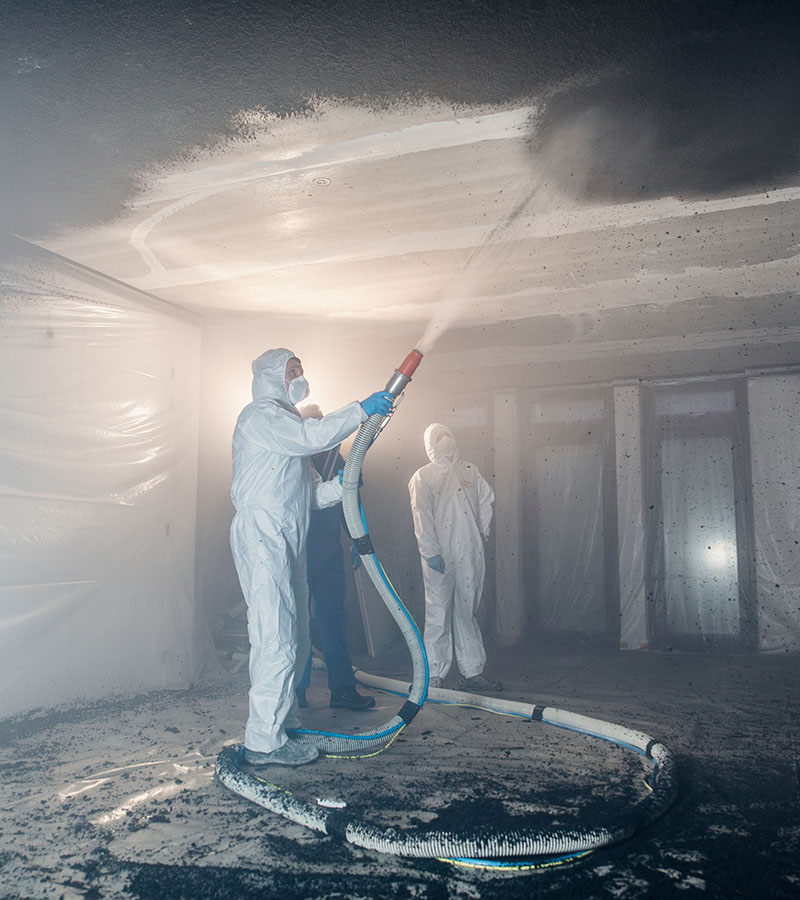 Personen in beschermende pakken en maskers brengen een akoestisch sprayplafond aan in een ruimte. Het proces genereert nevel terwijl het materiaal tegen het plafond wordt gespoten voor geluidsisolatie en demping.
